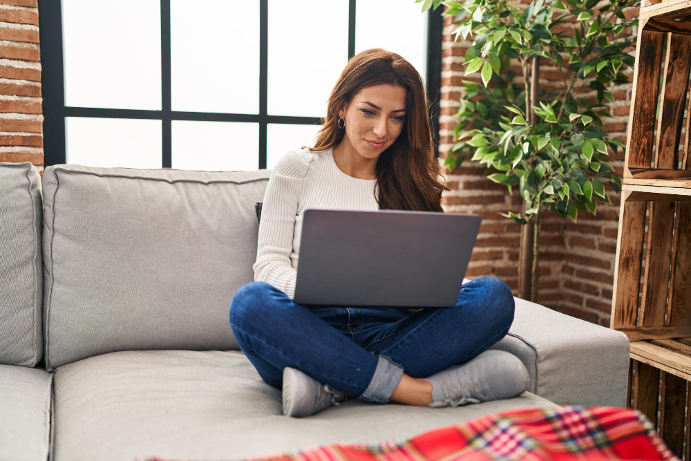 A woman using a laptop while sitting on the floor and leaning against a sofa.