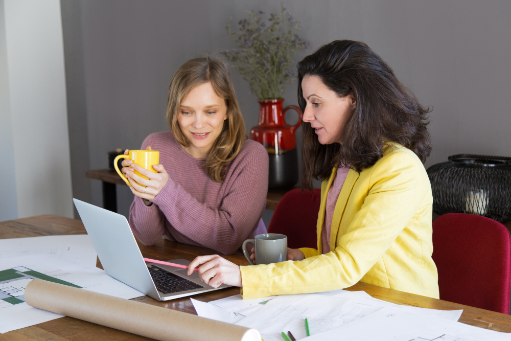 Two women sitting at a table with coffee cups while looking at a laptop.