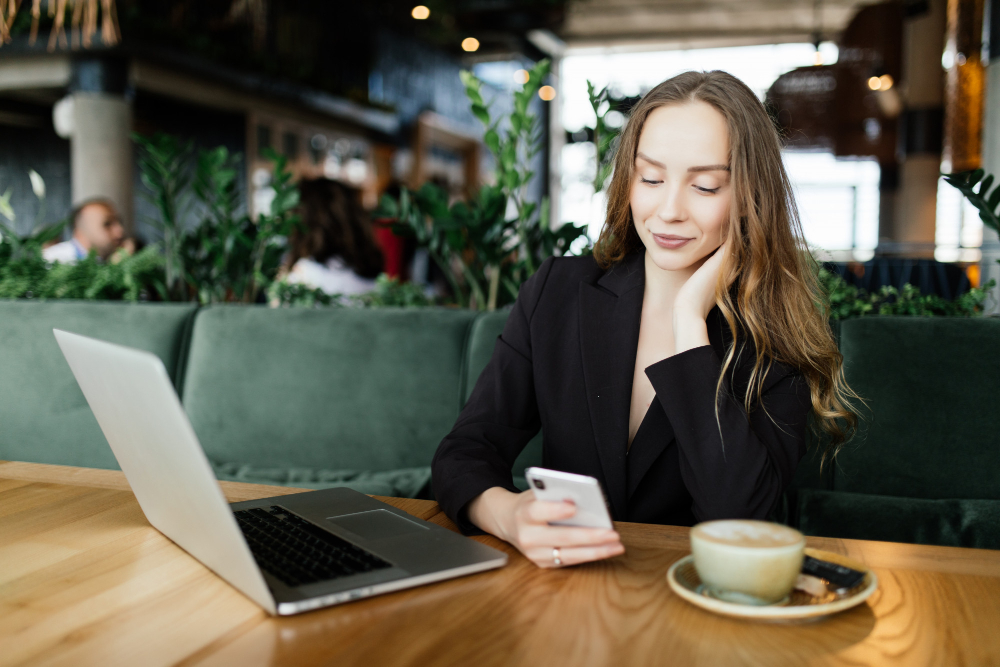 An image of a woman sitting in a cafe looking at her phone and her laptop.